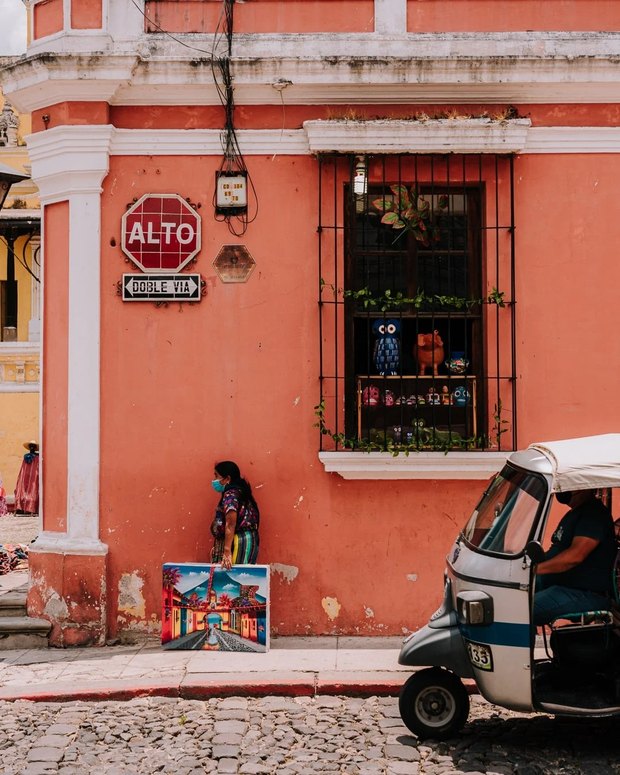 A coral-pink street corner with a barred window of painted wooden animals, a woman carrying a canvas, and a tuk-tuk on the cobbles.