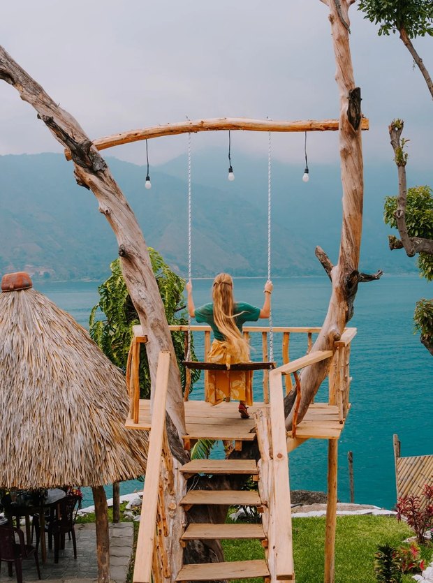 A rough timber swing hung from a branch on a small platform above the lake, with hills and turquoise water behind.