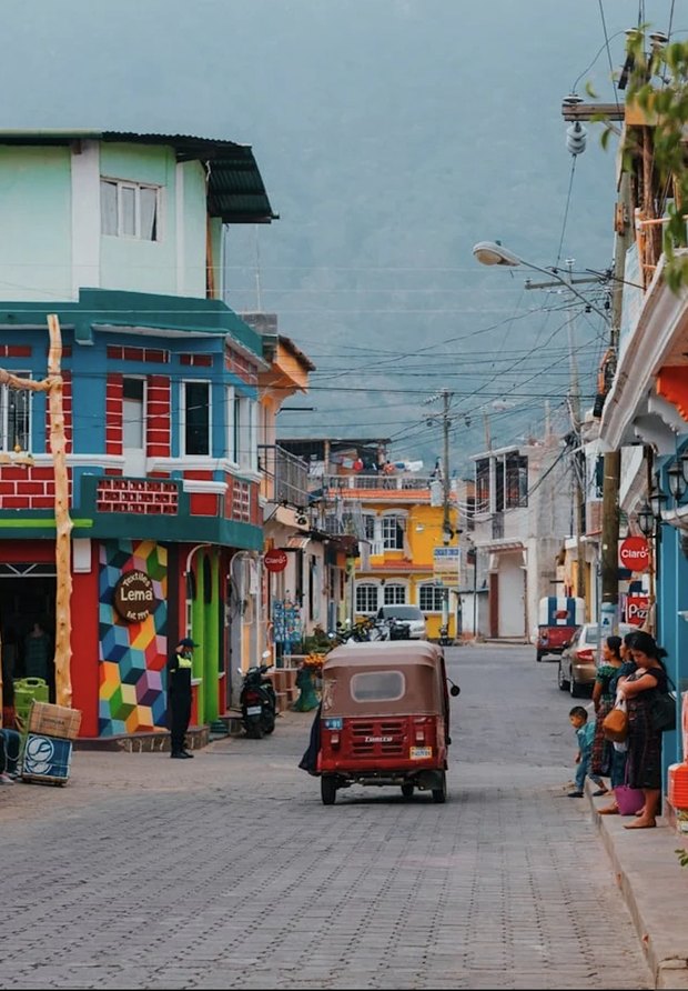 A village street of brightly painted buildings with a red tuk-tuk coming down the cobbles and the hillside in cloud behind.