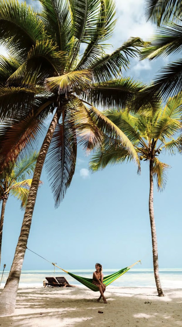 A green hammock slung between two palms on an empty beach, the sea flat and pale blue behind it.
