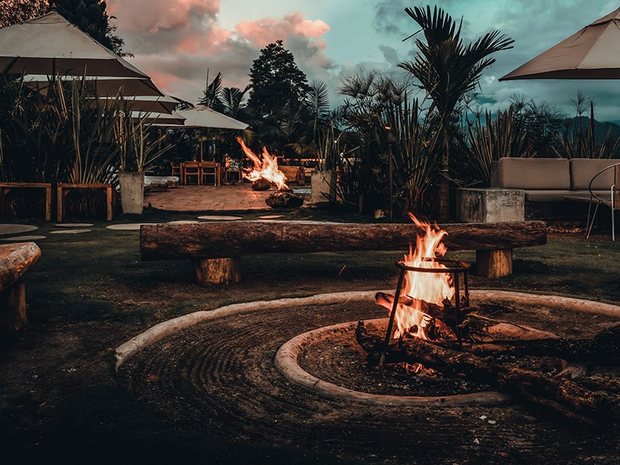 Fire pits burning in the garden at dusk, ringed by log benches and umbrellas under a pink sky.