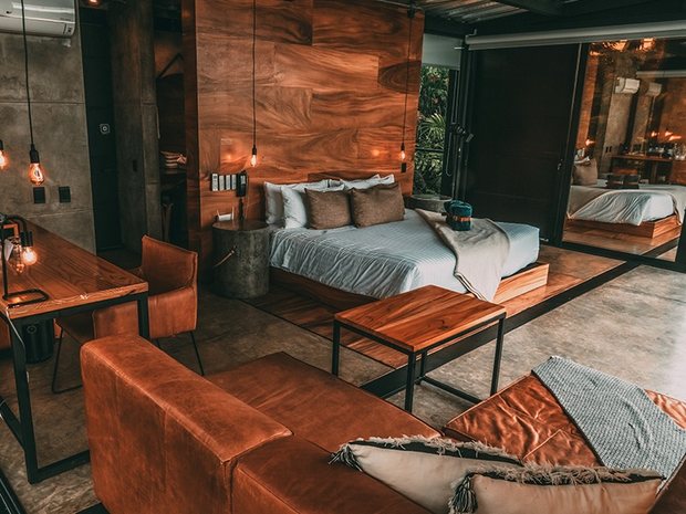 A suite interior in warm wood and concrete, with a tan leather sofa, a long desk and hanging bulbs.