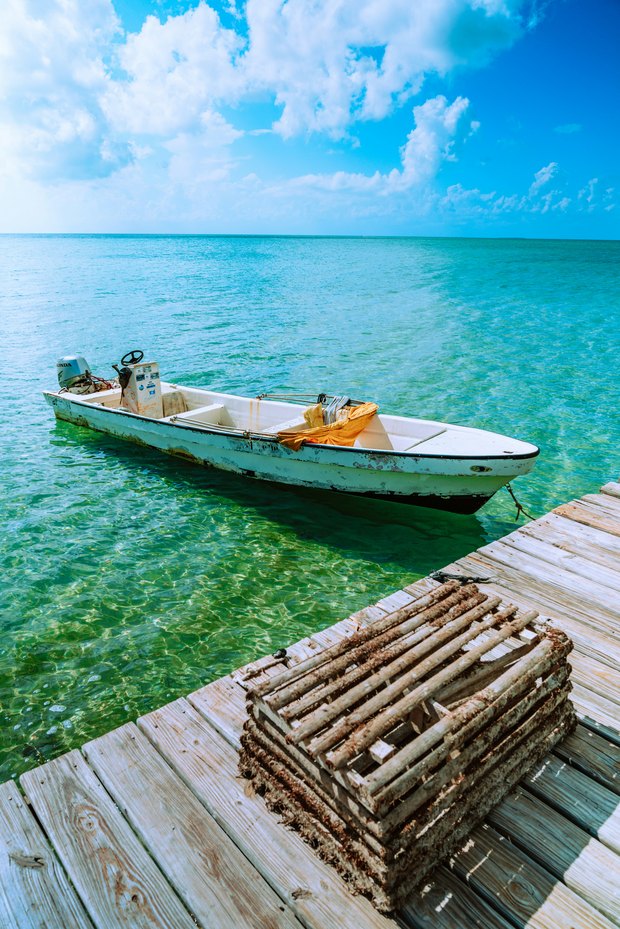 A small white fishing skiff moored off a weathered dock with a lobster trap on it, in shallow green-blue water.