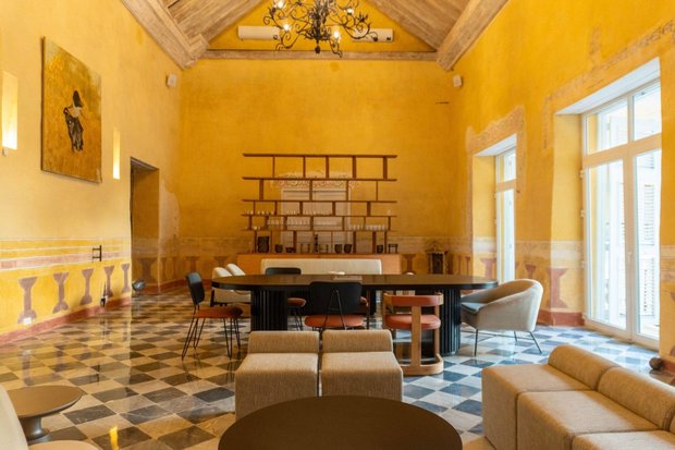 A tall yellow salon under bare timber beams and an iron chandelier, with restored wall murals and a chequerboard marble floor.