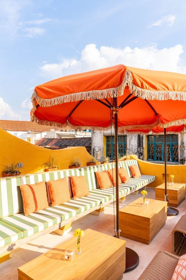 The roof terrace: a fringed orange parasol over striped banquettes, ochre walls, and the blue shutters of the old town behind.