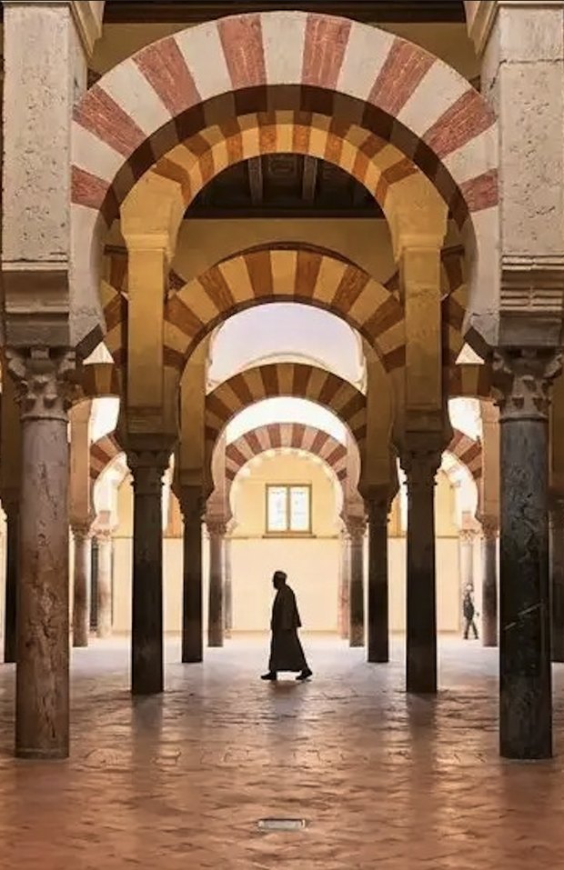 Inside the Mezquita: rows of red-and-white striped double arches on stone columns, a lone figure walking through.