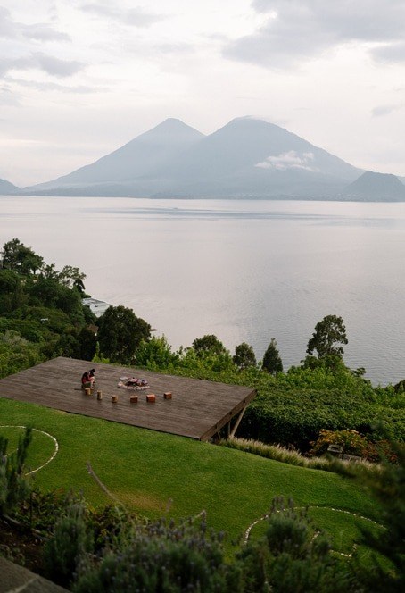 A timber deck on the lawn above Lake Atitlán at dusk, with two people sitting by a fire pit and two volcanoes across the water.