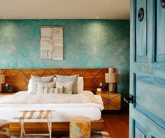 A room with a turquoise lime-washed wall, a wooden headboard, a woven wall hanging and a blue-painted door.