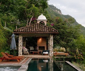 A stone pavilion with a tiled roof and a fireplace beside a dark pool, backed by jungle and a steep green hillside.