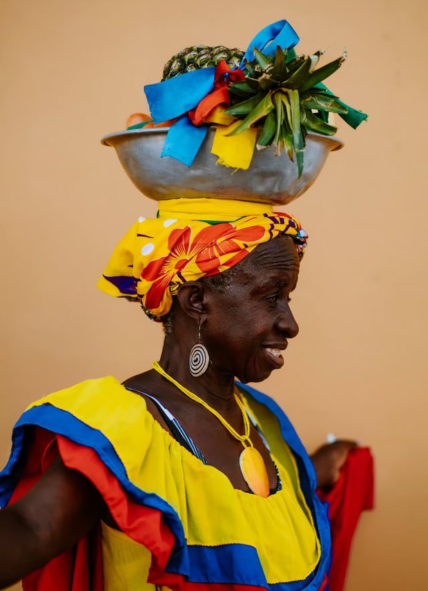 A palenquera in a yellow, blue and red ruffled dress, carrying a metal bowl of pineapples and mangoes balanced on her head.