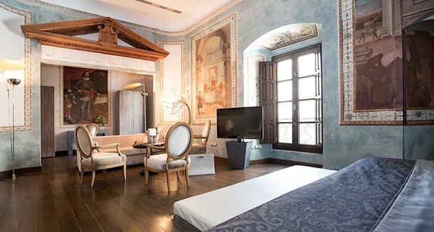 A room with blue-grey walls and old painted panels, a carved wooden pediment, velvet chairs and tall shuttered windows.