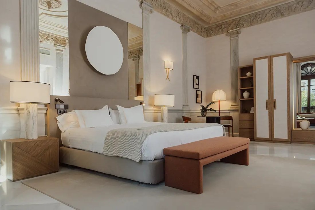A room at Palacio de los Patos: a wide bed under an ornate stucco ceiling, fluted columns, marble lamps and a terracotta bench, in soft cream and oak.