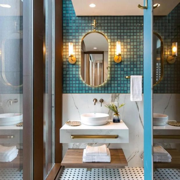 A bathroom in teal mosaic tile and marble, with a round basin, brass fittings and a mirror between two sconces.