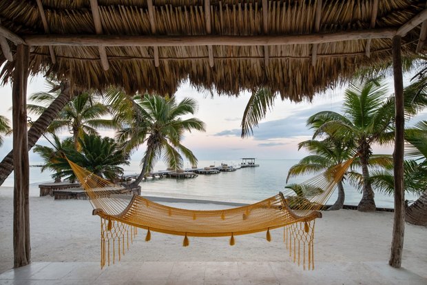 A yellow fringed hammock under a thatched roof, looking out through palms to the pier and a still sea at dawn.
