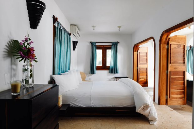 A white room with a low dark-wood bed, turquoise curtains, bougainvillea on the dresser and arched louvred wooden doors.