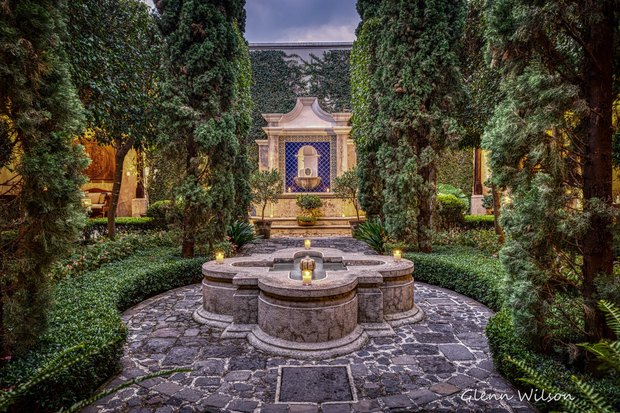The courtyard at dusk: a carved stone fountain on cobbles, cypresses and clipped hedges, and a tiled wall fountain lit by candles.