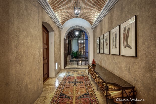 A vaulted brick corridor with lanterns, framed prints and a long table stacked with firewood, opening onto the courtyard.