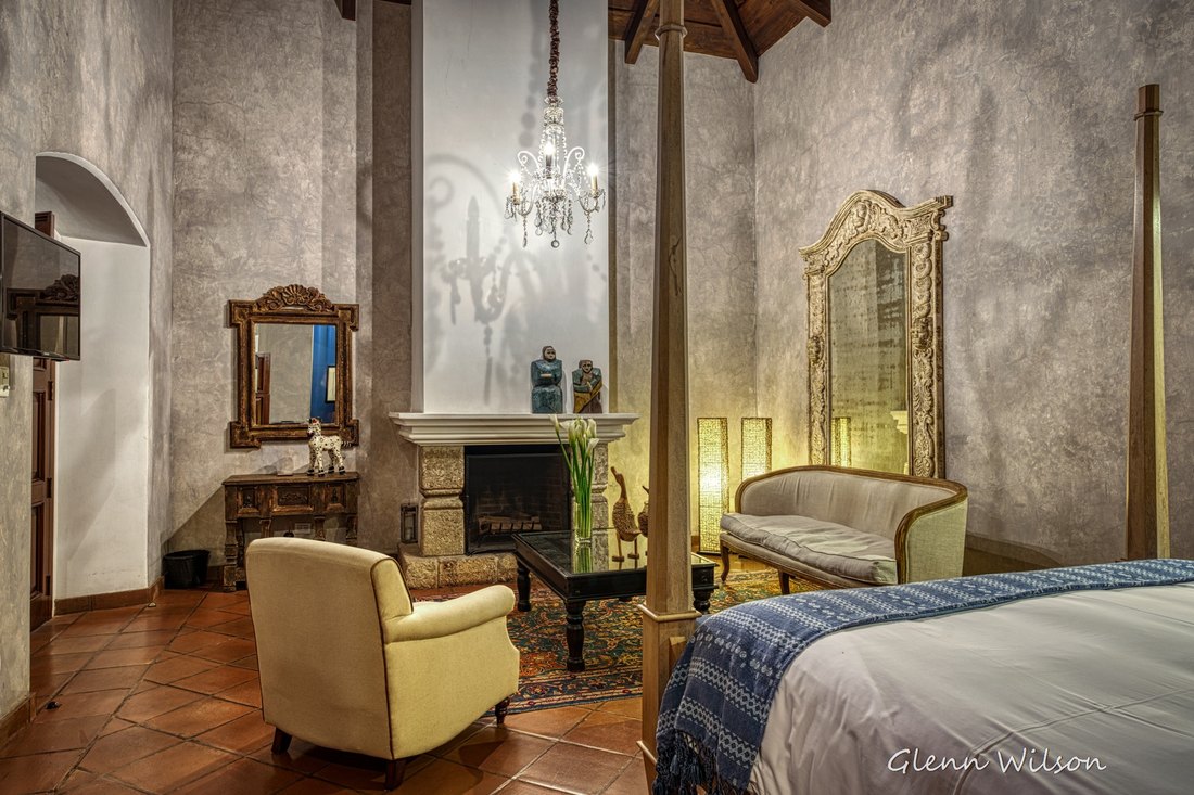 A room at San Rafael: a four-poster bed, a stone fireplace with a lit chandelier above it, a carved mirror and a sofa, under a high beamed ceiling.
