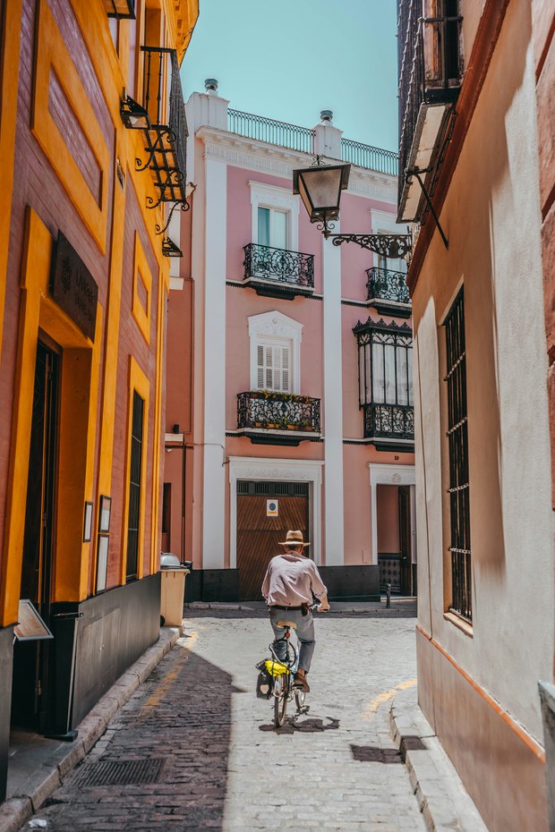 A narrow old-town street of ochre and pink houses with iron balconies, a man in a straw hat cycling down the cobbles.