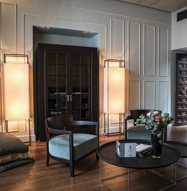 A sitting room with pale panelled walls, tall paper floor lamps, a dark glass-fronted cabinet and velvet armchairs.