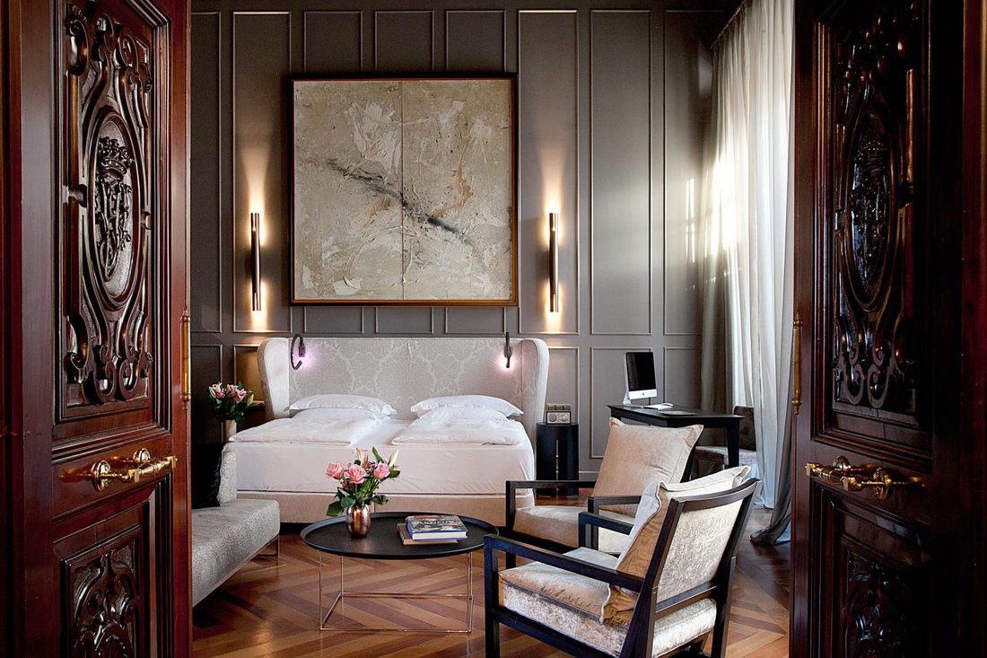 A room seen through carved mahogany doors: grey panelled walls, a large abstract painting over a white upholstered bed, velvet armchairs and herringbone floors.