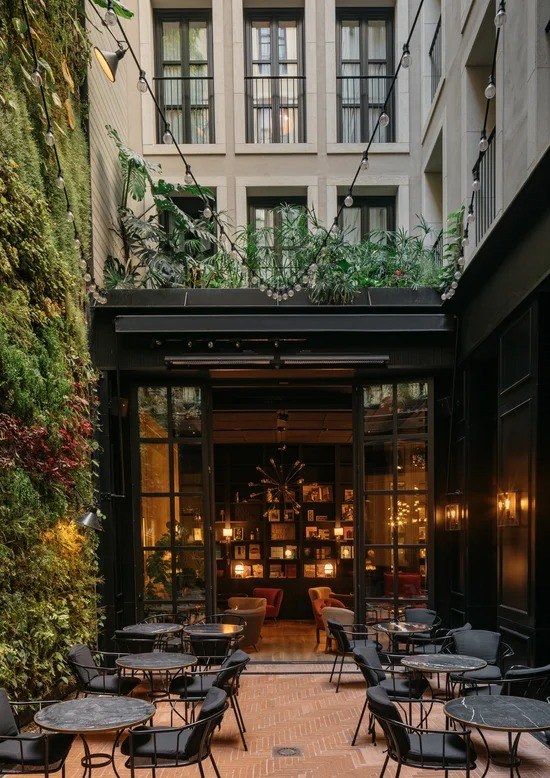 The courtyard: a tall living wall of plants, string lights and marble tables on terracotta tiles, the lit library visible through glass doors.